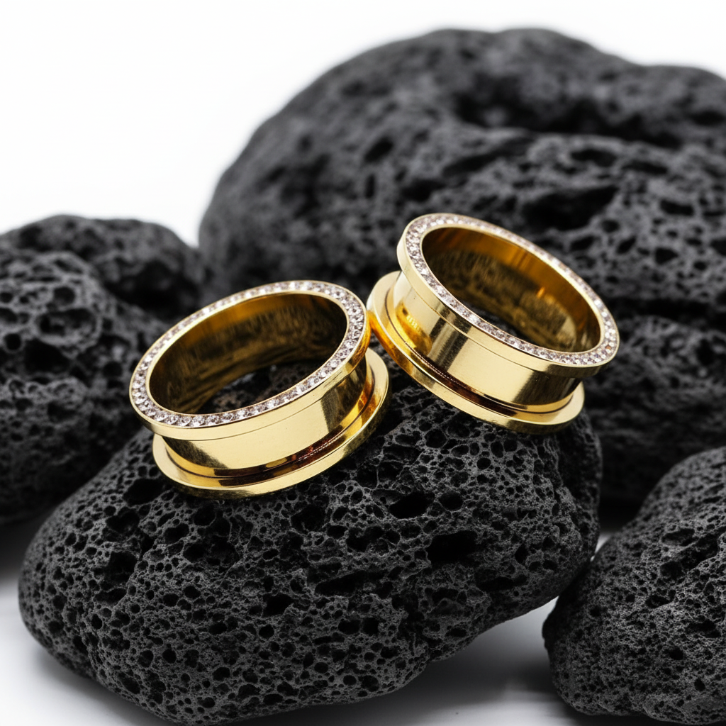 Pair of Gold PVD plated surgical steel tunnels lined with clear gems, shown resting on a dark surface. Available in sizes 2G up to 1 inch.