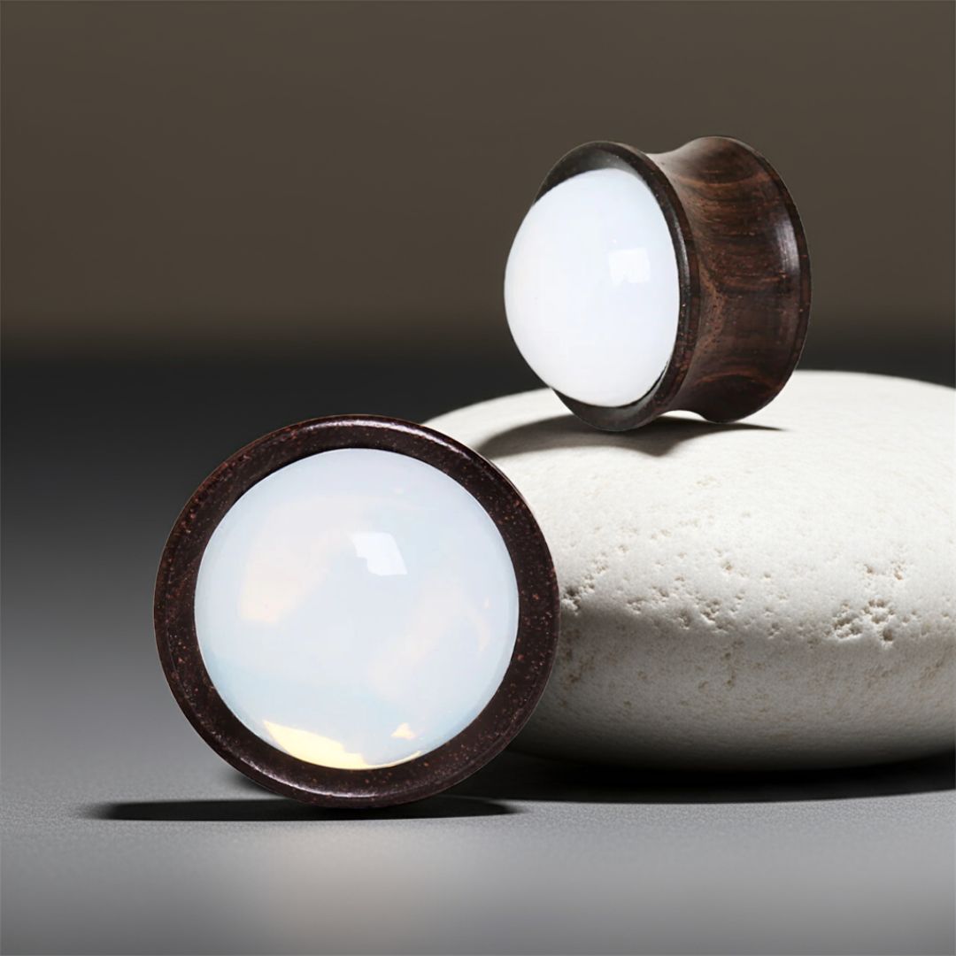 Pair of ebony wood ear plugs with shimmering opalite inlays, featuring rich dark grain and luminous center accents on a stone background
