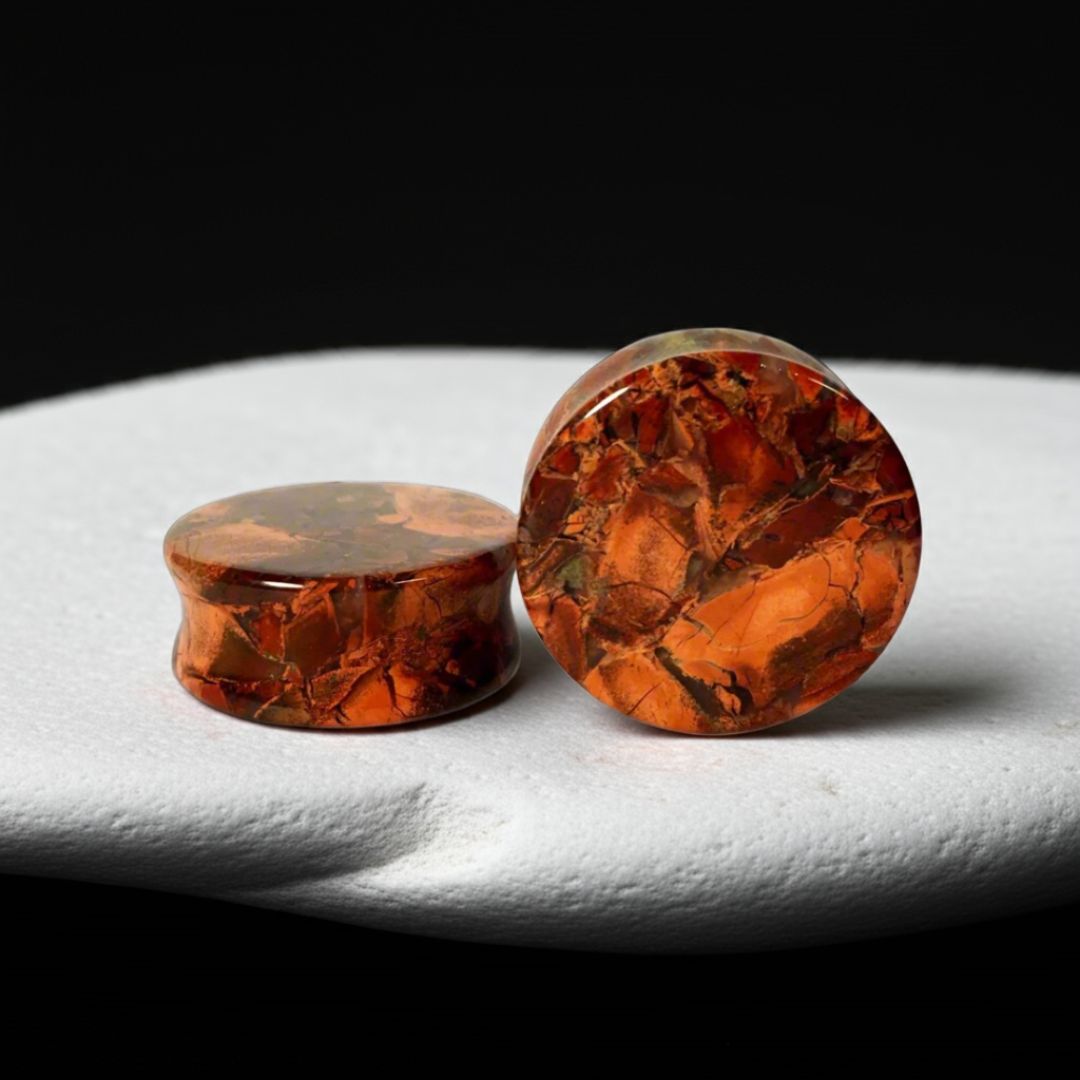Pair of poppy jasper stone ear plugs with deep red tones and natural earthy patterns, shown on a textured dark background