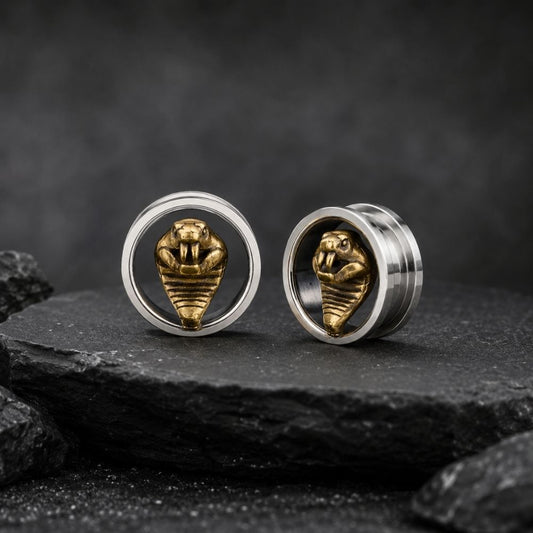 A pair of silver-toned flatback ear plugs with a gold-toned cobra snake design in the center, displayed on a dark textured slate surface.