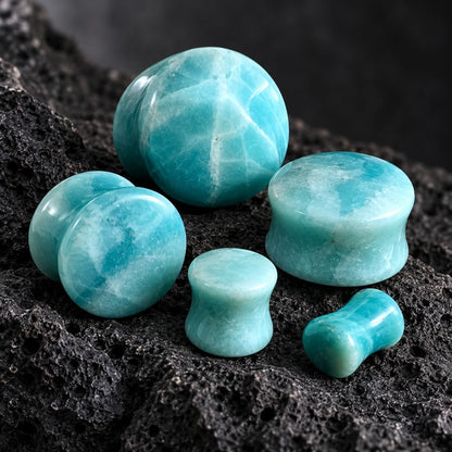 A set of turquoise-blue polished stone plugs and domes arranged on a dark porous rock surface. The pieces include round, cylindrical, and dumbbell-shaped forms with a marbled aqua coloration.