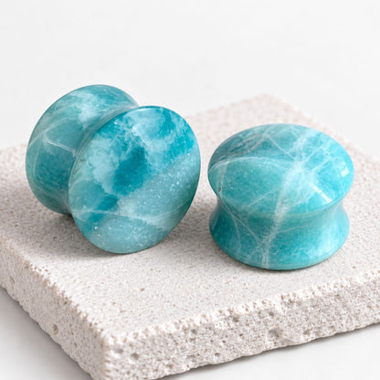 Two round turquoise-blue marbled plug earrings displayed on a textured light stone slab against a clean white background.
