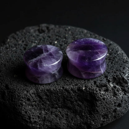 Pair of deep purple Amethyst stone plugs, double flare style, shown against a dark, textured background. Stretched ear jewelry.