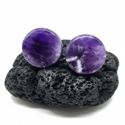 Pair of deep purple Amethyst stone plugs, double flare style, shown against a dark, textured background. Stretched ear jewelry.