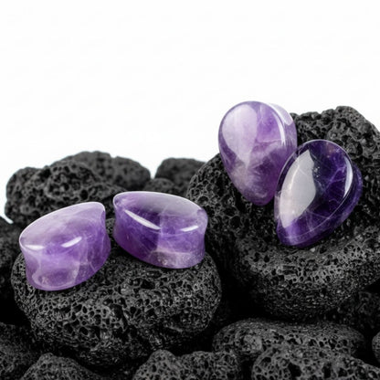 A close-up pair of deep purple, natural Amethyst stone teardrop plugs sitting on a dark surface. Stretched ear gauges jewelry.