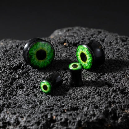 A pair of Basilisk Eye Acrylic Plugs (3mm-30mm) on dark volcanic rock with dramatic light.