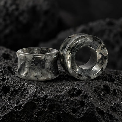 Two dark gray and black marbled stone ear plugs displayed on a textured black rock surface.