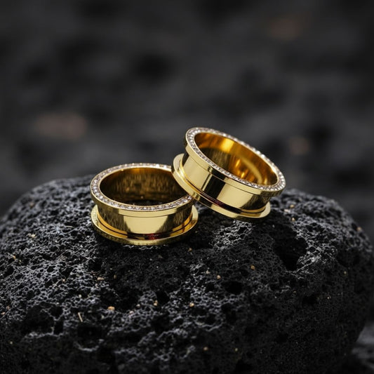 Pair of Gold PVD plated surgical steel tunnels lined with clear gems, shown resting on a dark surface. Available in sizes 2G up to 1 inch.