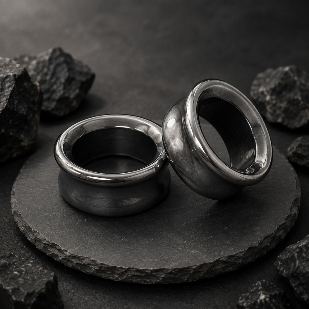 Two polished hematite plugs for stretched ears displayed on a round black stone slab, surrounded by rough dark rocks against a dark textured background.