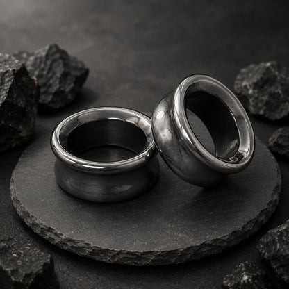 Two polished hematite plugs for stretched ears displayed on a round black stone slab, surrounded by rough dark rocks against a dark textured background.