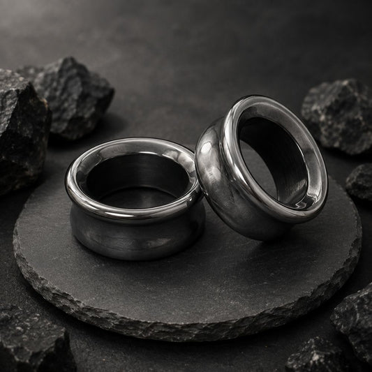 Two polished hematite plugs for stretched ears displayed on a round black stone slab, surrounded by rough dark rocks against a dark textured background.