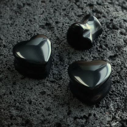 A pair of Onyx Stone Heart Plugs (6mm-16mm) resting on dark volcanic rock with sharp side lighting.