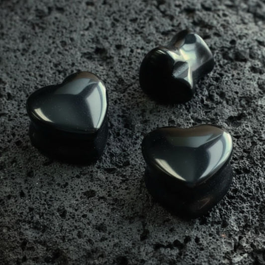 A pair of Onyx Stone Heart Plugs (6mm-16mm) resting on dark volcanic rock with sharp side lighting.