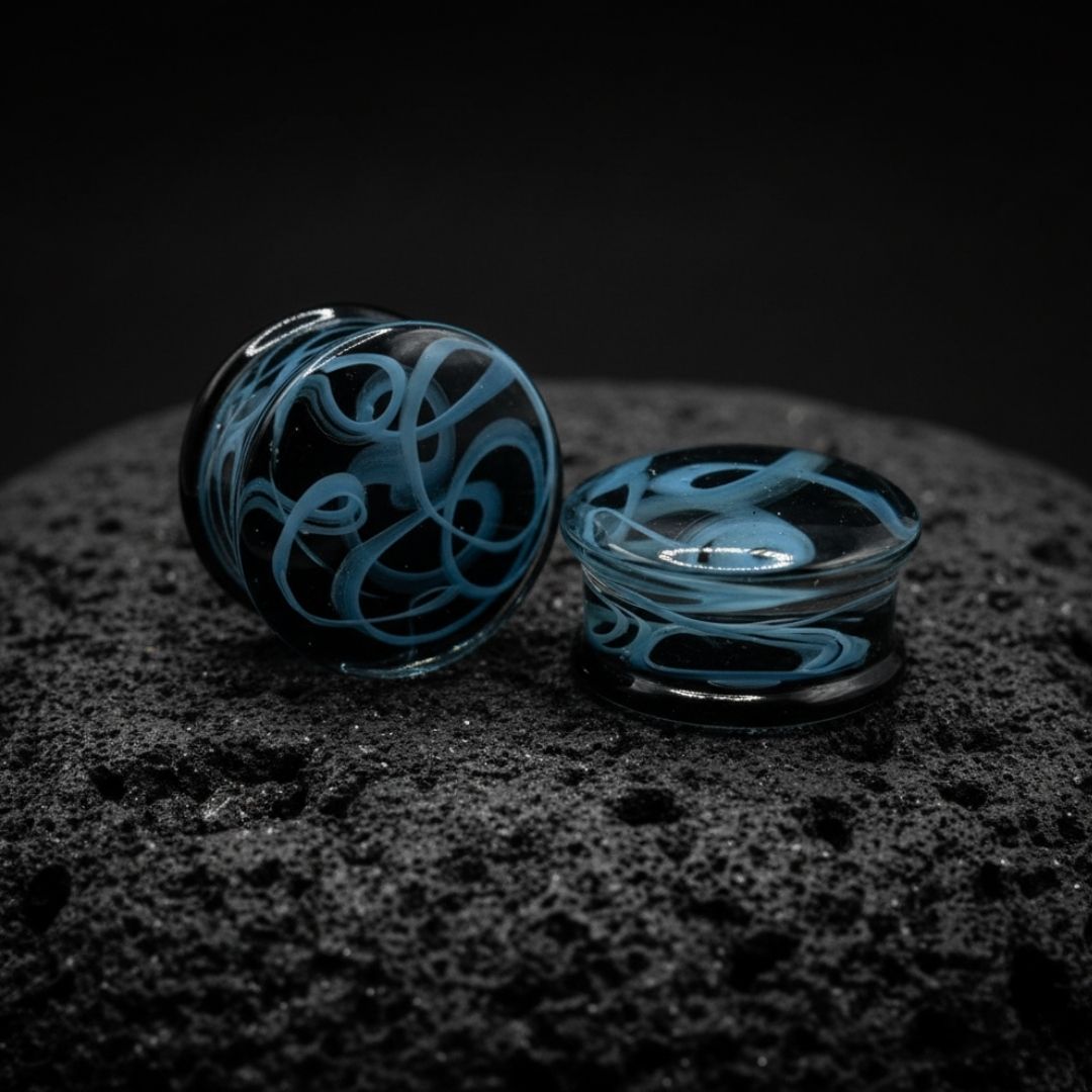 Pair of translucent glass plugs with dark smoky swirl patterns for stretched ear piercings, available in 6mm to 25mm.

