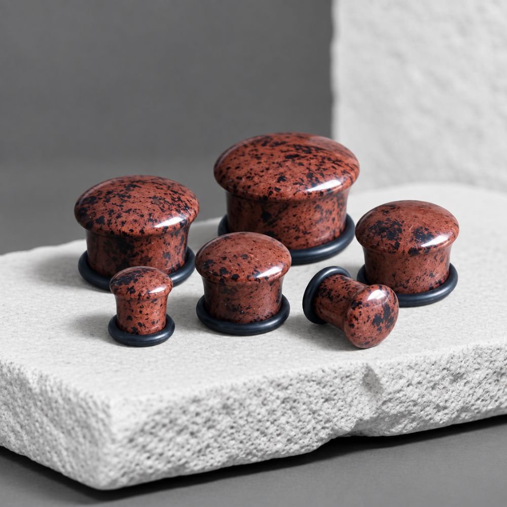 Six round mahogany obsidian gauges with a reddish-brown and black speckled granite-like finish, displayed on a textured light gray stone slab against a dark gray background.