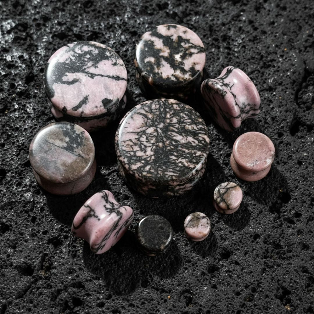 Rhodonite Stone Plugs (6mm-25mm) on dark volcanic rock with dramatic side lighting.