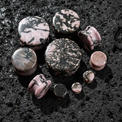 Rhodonite Stone Plugs (6mm-25mm) on dark volcanic rock with dramatic side lighting.
