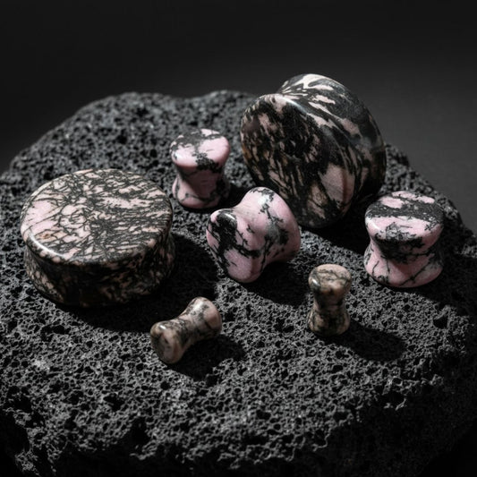 Rhodonite Stone Plugs (6mm-25mm) on dark volcanic rock with dramatic side lighting.