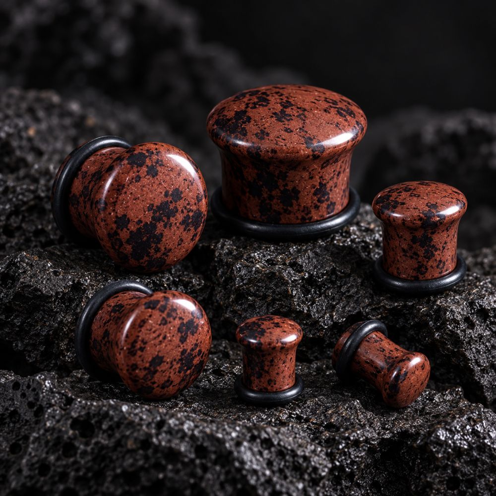 Six mahogany obsidian stone plug earrings with black speckles and black metal backing rings, arranged on a dark textured volcanic rock surface.