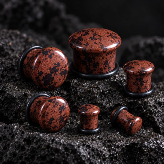 Six mahogany obsidian stone plug earrings with black speckles and black metal backing rings, arranged on a dark textured volcanic rock surface.