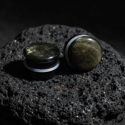 Single flare golden obsidian stone plugs shown up close on a black stone. Dark gauges with gold flashes, suitable for stretched ear piercings