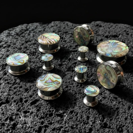 Steel Abalone Inlay Plugs (6mm-25mm) on dark volcanic rock with dramatic lighting.