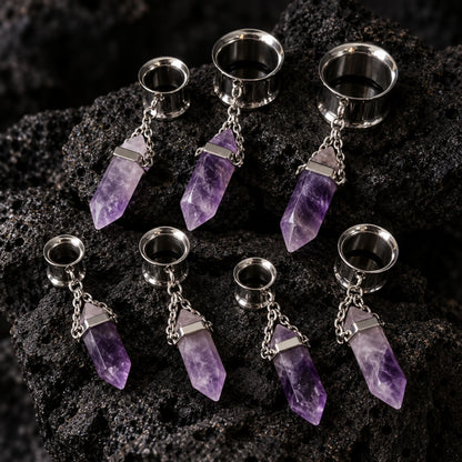 Steel Amethyst Dangle Tunnels (6mm-16mm) on dark volcanic rock with sharp shadows.