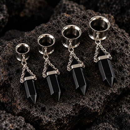 A pair of Steel Onyx Dangle Tunnels on dark volcanic rock with dramatic side lighting.