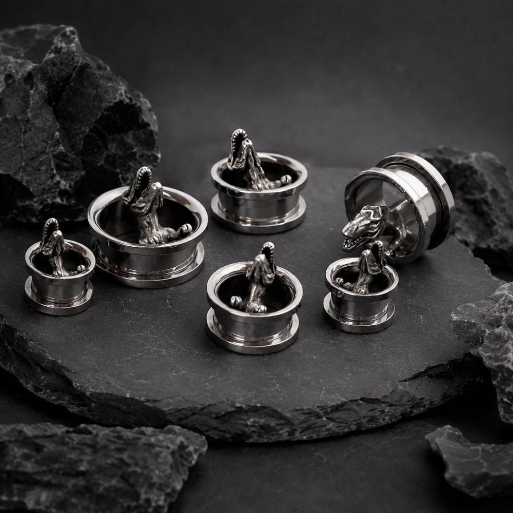 Six silver-toned metal ear plugs with detailed t rex carvings, arranged on a dark slate slab with black rocks around them.