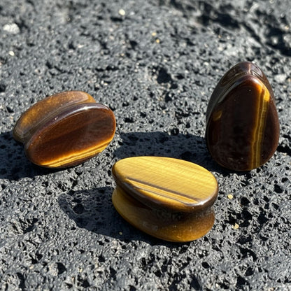 A pair of Tigers Eye Stone Teardrop Plugs (6mm-38mm) on dark volcanic rock with dramatic lighting.