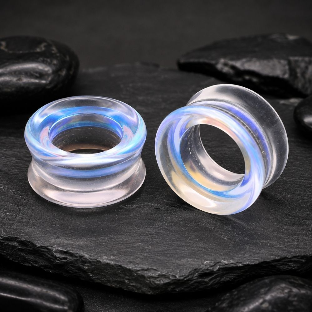 Two shimmer glass ear plugs with blue and white iridescent swirls, displayed on a dark slate surface with black stones in the background.